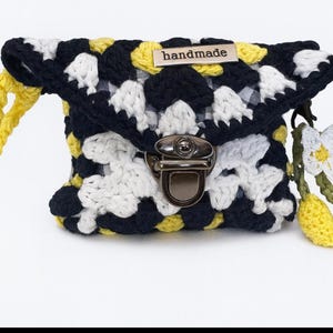 May include: Handmade crochet clutch in navy, white, and yellow. The bag features a metal clasp and a yellow handle. A small tag reads "handmade". A decorative charm with white flowers and yellow fruit hangs from the side.