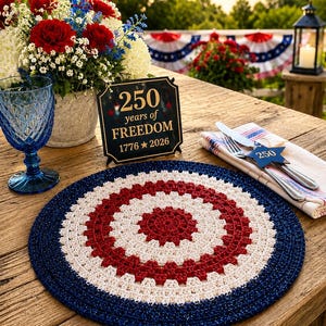 May include: A patriotic outdoor table setting with a red, white, and blue crochet placemat. A sign reads "250 years of FREEDOM 1776-2026". A bouquet of red and white flowers and a blue glass add to the festive decor.