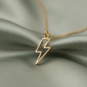Pode incluir: Um colar dourado com um pingente em forma de raio. O pingente tem um design vazado e a corrente é delicada. O colar é exibido em um tecido verde-sálvia macio, criando um fundo contrastante que destaca a joia.