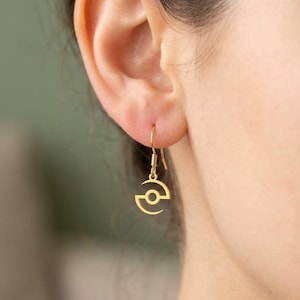 May include: A gold-toned drop earring featuring a circular charm with a central hole and two curved, crescent-like extensions on either side. The earring hangs from a simple gold-toned hook wire, suspended from the lower part of an earlobe.