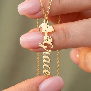 Puede incluir: Un collar dorado con un colgante de Snoopy y el nombre "Grace" en escritura vertical. El colgante está suspendido de una delicada cadena, sostenido entre dedos con esmalte de uñas rosa.