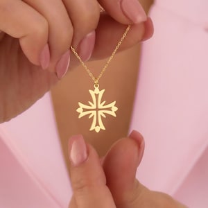 May include: A gold-colored pendant necklace featuring a decorative cross design. The cross has a symmetrical, openwork pattern with stylized floral or leaf-like elements at the ends of each arm. The necklace is held against a soft pink background.
