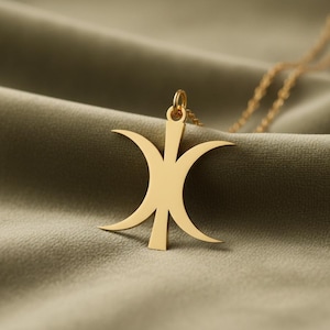 May include: A gold-colored pendant necklace with a unique, stylized design. The pendant features a central vertical bar with curved, crescent-shaped elements extending outwards. The necklace chain is also gold, and the background is a soft, olive-green fabric.