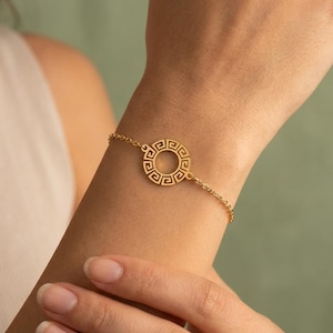 May include: A gold-colored bracelet featuring a circular pendant with a Greek key design. The bracelet is delicate, with a fine chain and a detailed, openwork pendant. The jewelry is displayed on a wrist against a soft green background.