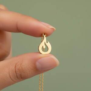 May include: A gold-colored pendant necklace featuring a flame design. The pendant is held between fingers against a soft green background. The necklace chain is also gold and delicate.