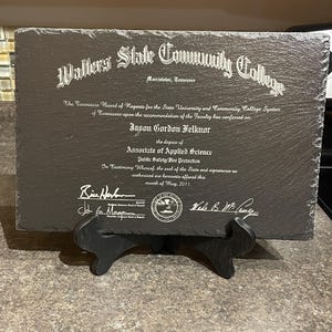 Puede incluir: Una placa de pizarra gris oscuro con texto grabado en blanco, exhibida sobre un soporte negro. La placa es un diploma del Walters State Community College, con el nombre de Jason Gordon Felknor y el t&iacute;tulo de Associate of Applied Science.