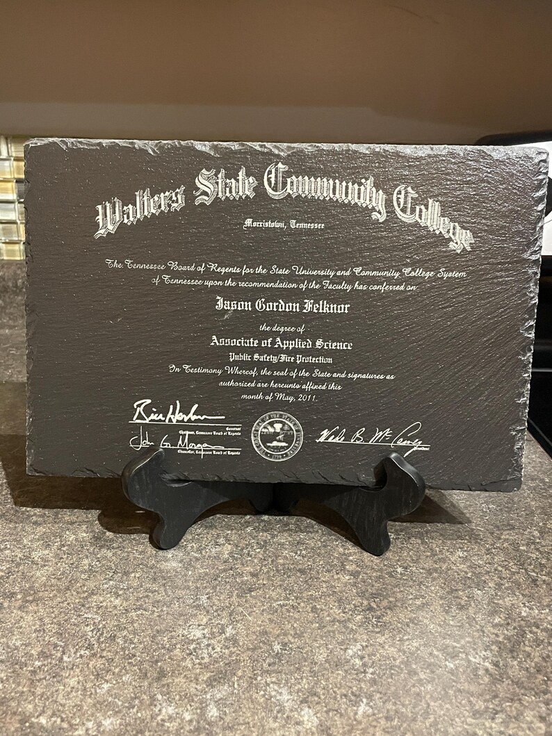 Puede incluir: Una placa de pizarra gris oscuro con texto grabado en blanco, exhibida sobre un soporte negro. La placa es un diploma del Walters State Community College, con el nombre de Jason Gordon Felknor y el t&iacute;tulo de Associate of Applied Science.