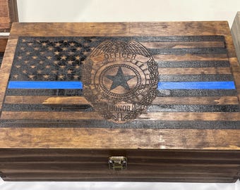 Puede incluir: Caja de madera con un diseño de bandera estadounidense desgastada, una línea azul y una insignia de policía. La caja tiene una tapa con bisagras y un cierre de metal. La caja está hecha de madera y tiene un acabado marrón oscuro.