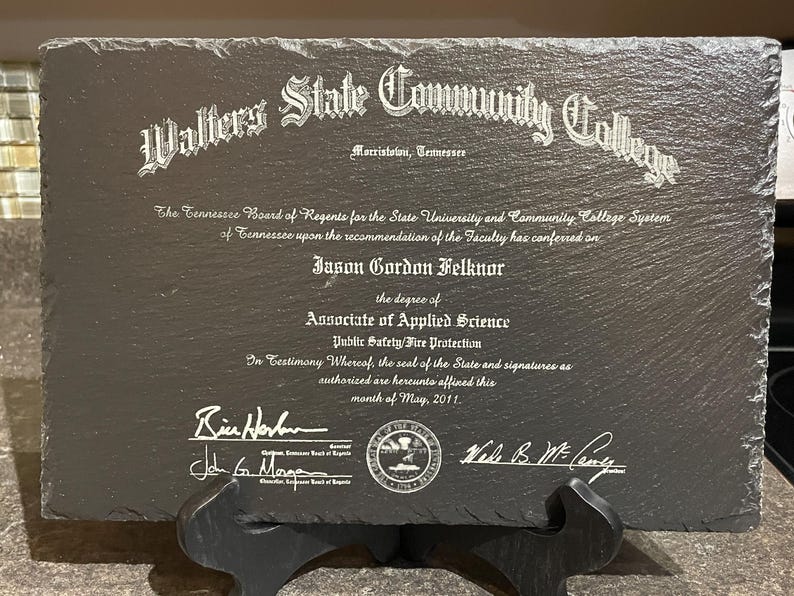Puede incluir: Una placa de pizarra gris oscuro con texto blanco grabado. El texto dice "Walters State Community College" e incluye un diploma para Jason Gordon Felknor, con el t&iacute;tulo de Associate of Applied Science. La placa se muestra en un soporte negro.