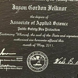 Puede incluir: Placa de pizarra negra con texto blanco grabado. El texto dice "Jason Gordon Felknor" y detalla un t&iacute;tulo de Asociado en Ciencias Aplicadas en Seguridad P&uacute;blica/Protecci&oacute;n contra Incendios. Incluye firmas, un sello estatal y la fecha de mayo de 2011.