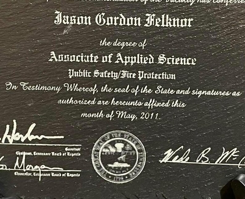 Puede incluir: Placa de pizarra negra con texto blanco grabado. El texto dice "Jason Gordon Felknor" y detalla un t&iacute;tulo de Asociado en Ciencias Aplicadas en Seguridad P&uacute;blica/Protecci&oacute;n contra Incendios. Incluye firmas, un sello estatal y la fecha de mayo de 2011.