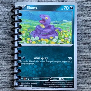 May include: A spiral-bound notebook featuring a Pokémon trading card. The card displays a purple Ekans snake character with the text "Ekans" and "Acid Spray". The card is set against a gray, textured background.