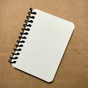 Puede incluir: Un cuaderno blanco de espiral con una encuadernación en espiral negra. El cuaderno está acostado sobre una superficie marrón.
