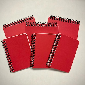 May include: A collection of six small, red spiral-bound notebooks. Each notebook has a black spiral binding and a solid red cover. The notebooks are arranged in a slightly overlapping pattern against a white background.