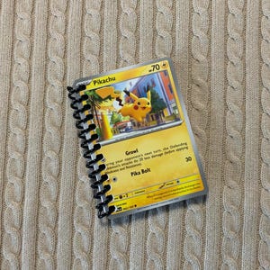 Może przedstawiać: Żółta karta Pokemon Pikachu z kreskówkowym obrazkiem Pikachu z przodu. Karta jest częścią spiralnie oprawionego notesu.