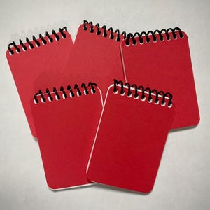 May include: Five small, red spiral-bound notebooks are arranged on a white surface. Each notebook has a black spiral binding and a bright red cover. The notebooks are rectangular with rounded corners.
