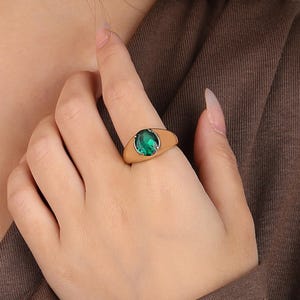 Op de afbeelding: Een goudkleurige ring met een grote, ovaalvormige, smaragdgroene edelsteen. De ring heeft een eenvoudige, gladde band. De ring is een sieraad.