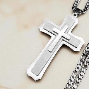 May include: A silver-colored cross pendant necklace. The cross has a layered design with a smaller cross in the center. The chain is a curb style. The necklace is displayed on a light-colored surface.