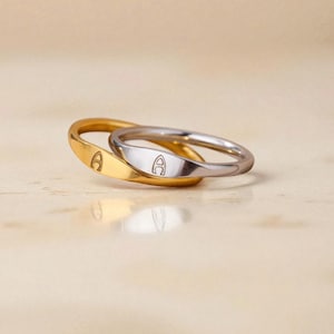 Puede incluir: Dos anillos, uno dorado y otro plateado, entrelazados. Cada anillo presenta una pequeña letra "A" grabada en su superficie. Los anillos se encuentran sobre un fondo neutro de color claro, creando una estética sencilla y elegante.