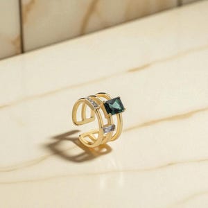 May include: A gold-coloured ring with an open design, featuring a square, dark green gemstone and smaller, clear stones. The ring has a multi-band design and is set against a light-coloured, marble-like surface.