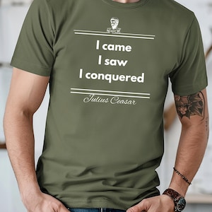 Puede incluir: Camiseta verde oliva con la cita "Vine, vi, vencí" atribuida a Julio César. El diseño incluye un busto de César encima de la cita. La camiseta es de manga corta y cuello redondo.