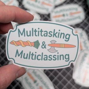 May include: A white sticker with a green border and rounded edges. The sticker features a stylized illustration of a sword and an axe crossed, with the text "Multitasking & Multiclassing" in green.