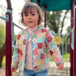 May include: A young child wearing a colourful patchwork jacket with a pink turtleneck sweater and blue jeans. The jacket has a variety of floral and geometric patterns in shades of red, pink, blue, green and white.