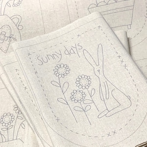 May include: A stack of light beige linen napkins with a hand-stitched design. Each napkin features a gray outline of a bunny, sunflowers, and the words "Sunny days". The design is enclosed within a dotted oval.