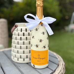 May include: A champagne bottle with a yellow and black bee design, a bright orange label, and a white bow. The label reads "Veuve Clicquot Brut". The bottle is on a gray wooden table next to a white lantern.