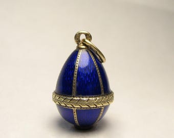 Puede incluir: Un colgante de huevo de esmalte azul con detalles dorados. El huevo está decorado con una banda dorada y un bucle dorado en la parte superior para colgar.