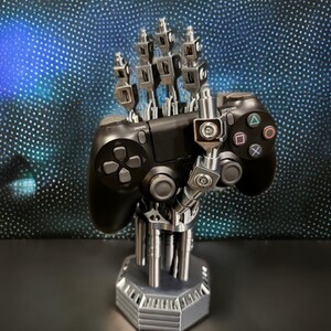 以下が含まれることがあります： 黒いビデオゲームコントローラーを保持する銀色の機械的なハンドスタンド。スタンドは関節のある金属のような指と六角形のベースを備えています。コントローラーには、標準的な方向ボタン、アナログスティック、アクションシンボルが含まれています。