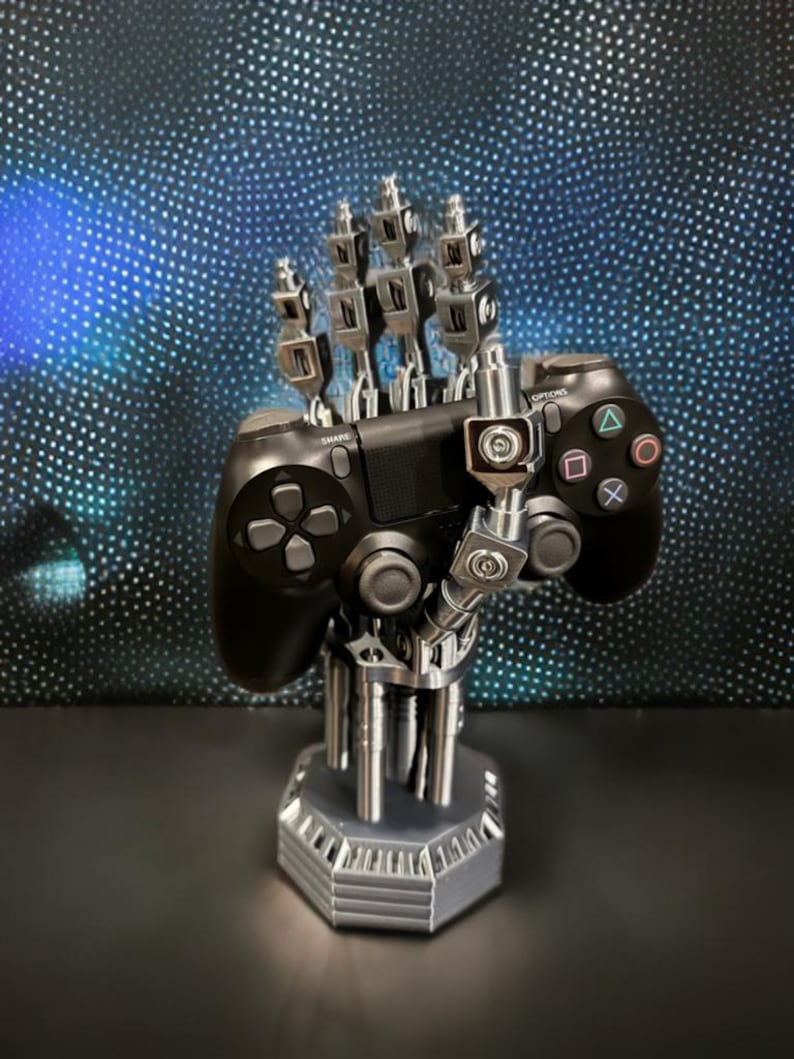以下が含まれることがあります： 黒いビデオゲームコントローラーを保持する銀色の機械的なハンドスタンド。スタンドは関節のある金属のような指と六角形のベースを備えています。コントローラーには、標準的な方向ボタン、アナログスティック、アクションシンボルが含まれています。