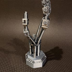以下が含まれることがあります： 銀色のロボットハンドの彫刻。関節のある機械的な指とジョイントを備え、頑丈な六角形の台座に取り付けられています。複雑な金属部品、目に見えるネジ、そして手の解剖学的構造を模したセグメント化されたロッドで構成されています。