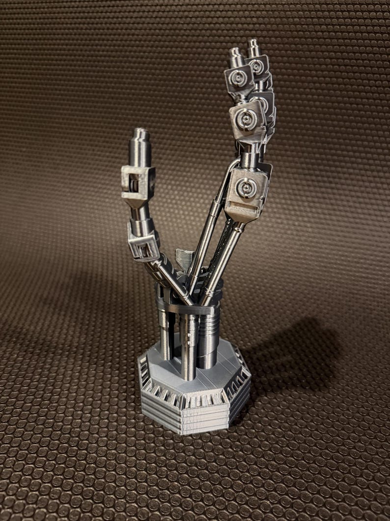 以下が含まれることがあります： 銀色のロボットハンドの彫刻。関節のある機械的な指とジョイントを備え、頑丈な六角形の台座に取り付けられています。複雑な金属部品、目に見えるネジ、そして手の解剖学的構造を模したセグメント化されたロッドで構成されています。