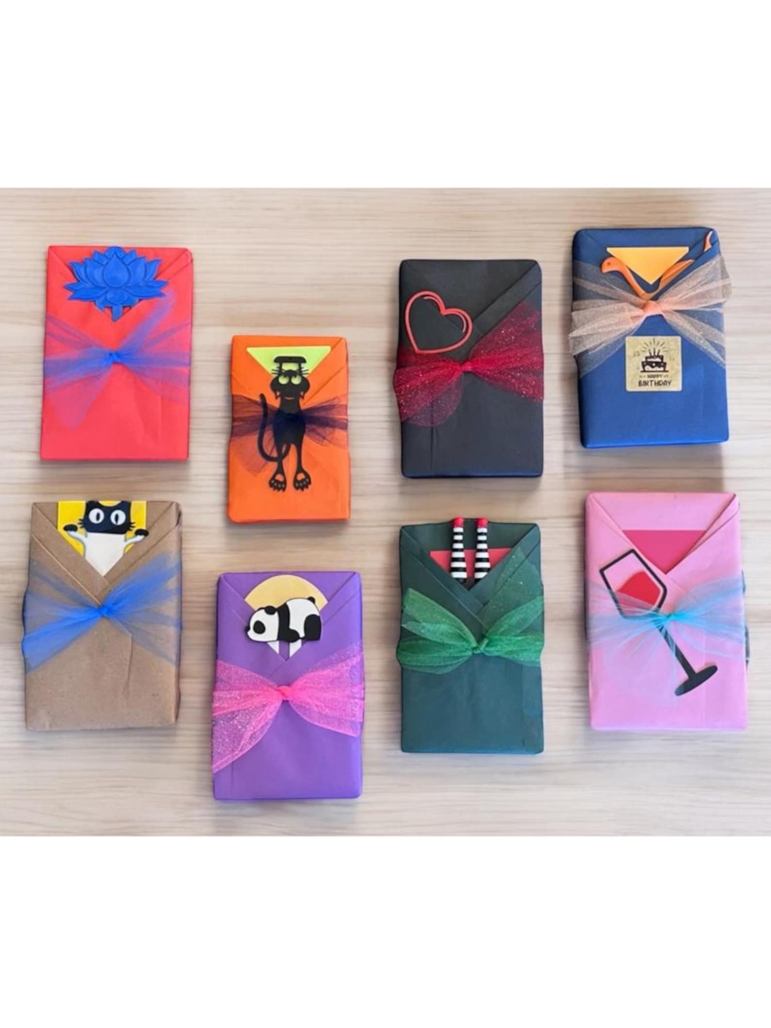 Books wrapped in wrapping paper of various different colors are laid against a hardwood floor. The top row features, from left to right: a book wrapped in red wrapping paper with blue detail, one wrapped in orange with black detail and a cat decal on it, one wrapped in black wrapping paper with red detail and a heart detail, and one wrapped in blue with a "happy birthday" sticker and light pink detail. On the bottom row, from left to right: a book wrapped in brown paper with a blue bow on it and a black cat wearing a white turtleneck illustrated on top, one wrapped in purple with a panda illustration and hot pink bow wrapped underneath it, one wrapped in dark green with a dark green bow and an illustration of two legs wearing black and white socks and red shoes sticking outward, and one wrapped in pink paper with a light blue ribbon and an illustration of a wine glass.