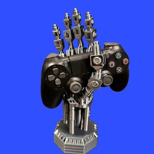 May include: A silver-colored robotic hand stand designed to hold a black video game controller. The mechanical hand features articulated fingers and is mounted on a sturdy, hexagonal base with vertical support rods, providing a stable display.