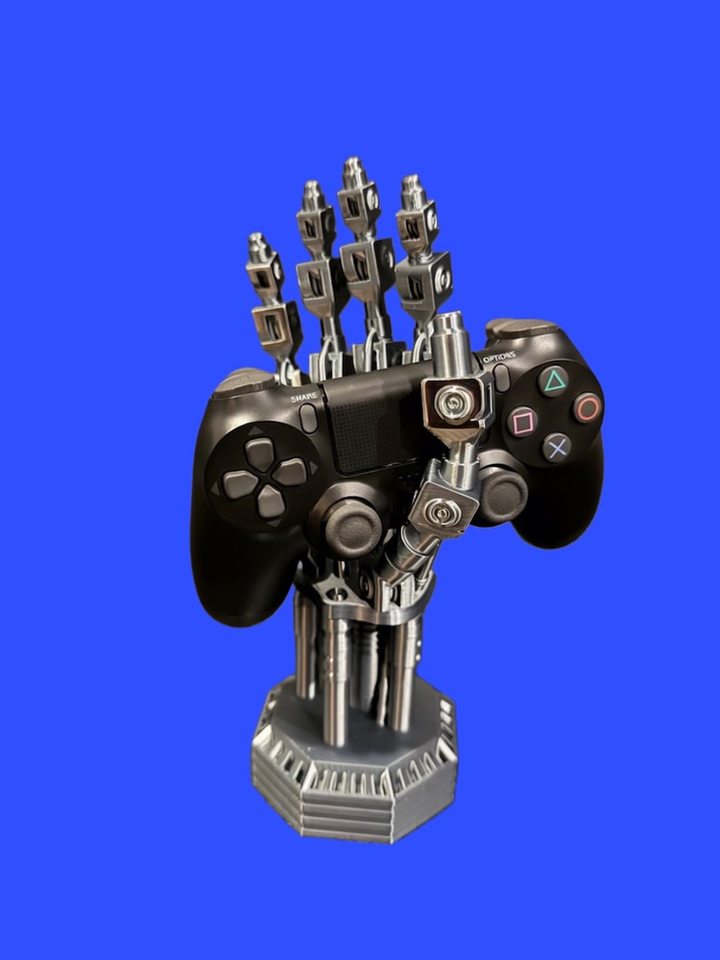 以下が含まれることがあります： 黒いビデオゲームコントローラーを保持するように設計された、銀色のロボットハンド型スタンド。機械的な手には関節のある指があり、垂直の支柱を備えた頑丈な六角形のベースに取り付けられており、安定したディスプレイを提供します。
