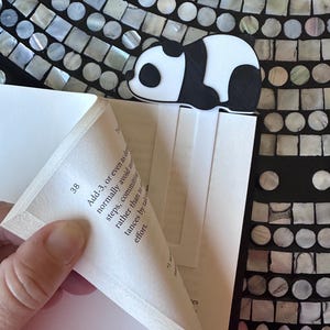 May include: A black and white panda bookmark resting on the pages of a book. The panda is 3D printed and has a cute design. The book is open, and the pages are white. The background is a mosaic of iridescent tiles.