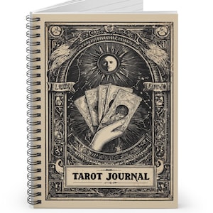 Può includere: Tarot Journal rilegato a spirale con copertina beige. La copertina presenta una dettagliata illustrazione in bianco e nero di una mano che tiene carte dei tarocchi, un sole e le parole "Tarot Journal". Il diario ha pagine a righe.