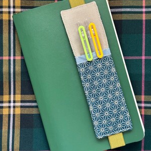 Op de afbeelding: Een groen notitieboek met een stoffen pennenhouder en twee pennen. De pennenhouder is beige met een blauw en wit geometrisch patroon. De pennen zijn groen en geel. Een gele elastische band houdt de pennenhouder aan het notitieboek.