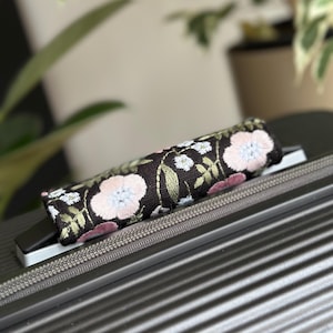 May include: A black luggage handle wrap featuring embroidered pink and white flowers and green leaves. The handle wrap is placed on a grey suitcase with a zip.