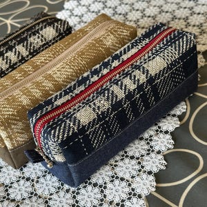 Può includere: Tre astucci rettangolari con chiusure a cerniera. L'astuccio più in primo piano è blu navy con una cerniera rossa e un motivo a quadri bianchi e blu navy. Gli altri due astucci sono beige e neri con motivi e cerniere simili.