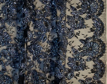 May include: Close-up of black lace fabric with a floral design. The flowers are outlined with a shiny, silver-blue material, creating a textured and dimensional effect. The lace has a delicate, open weave, and the edges are finished with a scalloped trim.