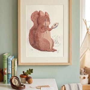May include: A framed watercolour print featuring a brown squirrel wearing round spectacles and holding a compass. The artwork is set in a light wood frame and includes the text "DOLCE FANZIA" in a soft, muted font on the right side of the illustration.