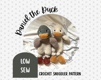 May include: Two crocheted duck toys, one with a green head and the other with a cream-colored head, are featured in this image. The ducks have orange beaks and feet, and brown wings. The text "Daniel the Duck" and "Crochet Snuggler Pattern" are also visible.