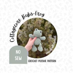 May include: A light green crocheted frog plushie with black eyes and a smiling expression. The image includes the text "Cottagecore Bubs-Frog" and "NO SEW CROCHET PLUSHIE PATTERN". The frog is held in a hand.