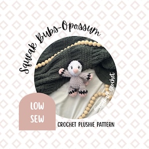 May include: A crocheted plushie opossum in gray, white, and pink, with black accents on its paws. The text "Squeak Bubs-Opossum" and "Crochet Plushie Pattern" are visible. The plushie is set against a dark green blanket and wooden beads.