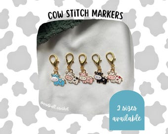 May include: A collection of five cow stitch markers with gold-colored clasps. Each marker features a different colored cow design: blue, white, pink, black, and red. The text "COW STITCH MARKERS" is at the top, and "2 sizes available" is in a blue box.