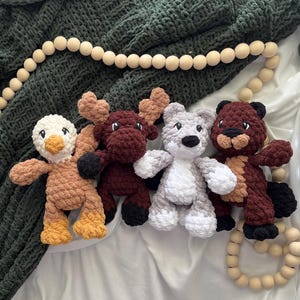 May include: Four handmade crochet animal toys: a duck, a moose, a bear, and a beaver. The duck is tan and yellow, the moose is burgundy, the bear is gray and white, and the beaver is brown. They are arranged on a white surface with a green blanket.