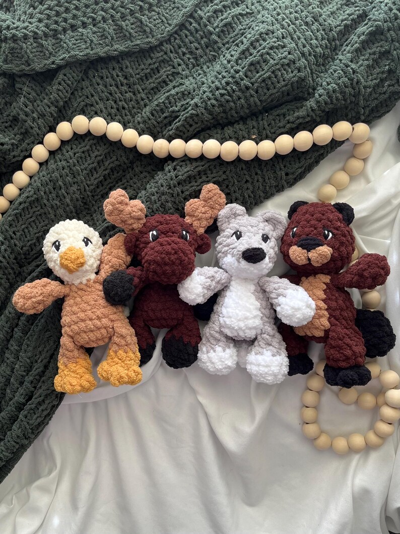 May include: Four handmade crochet animal toys: a duck, a moose, a bear, and a beaver. The duck is tan and yellow, the moose is burgundy, the bear is gray and white, and the beaver is brown. They are arranged on a white surface with a green blanket.